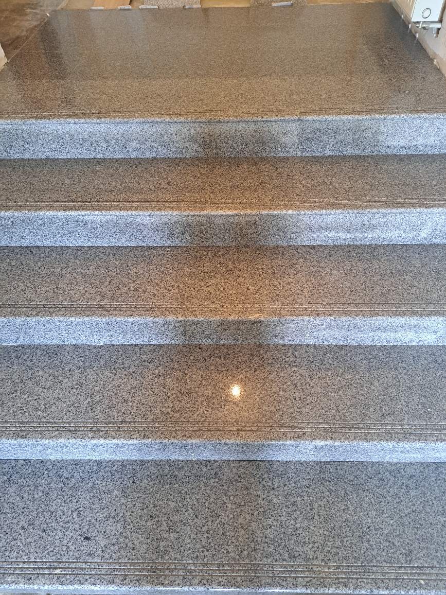 Schody wyłożone szarym, nakrapianym granitem, z widocznymi liniami antypoślizgowymi na krawędziach stopni.