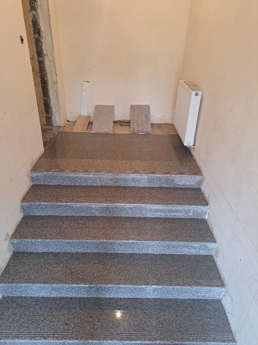 Schody wyłożone szarym kamieniem, z widocznym grzejnikiem i przygotowanymi płytkami do dalszych prac wykończeniowych.