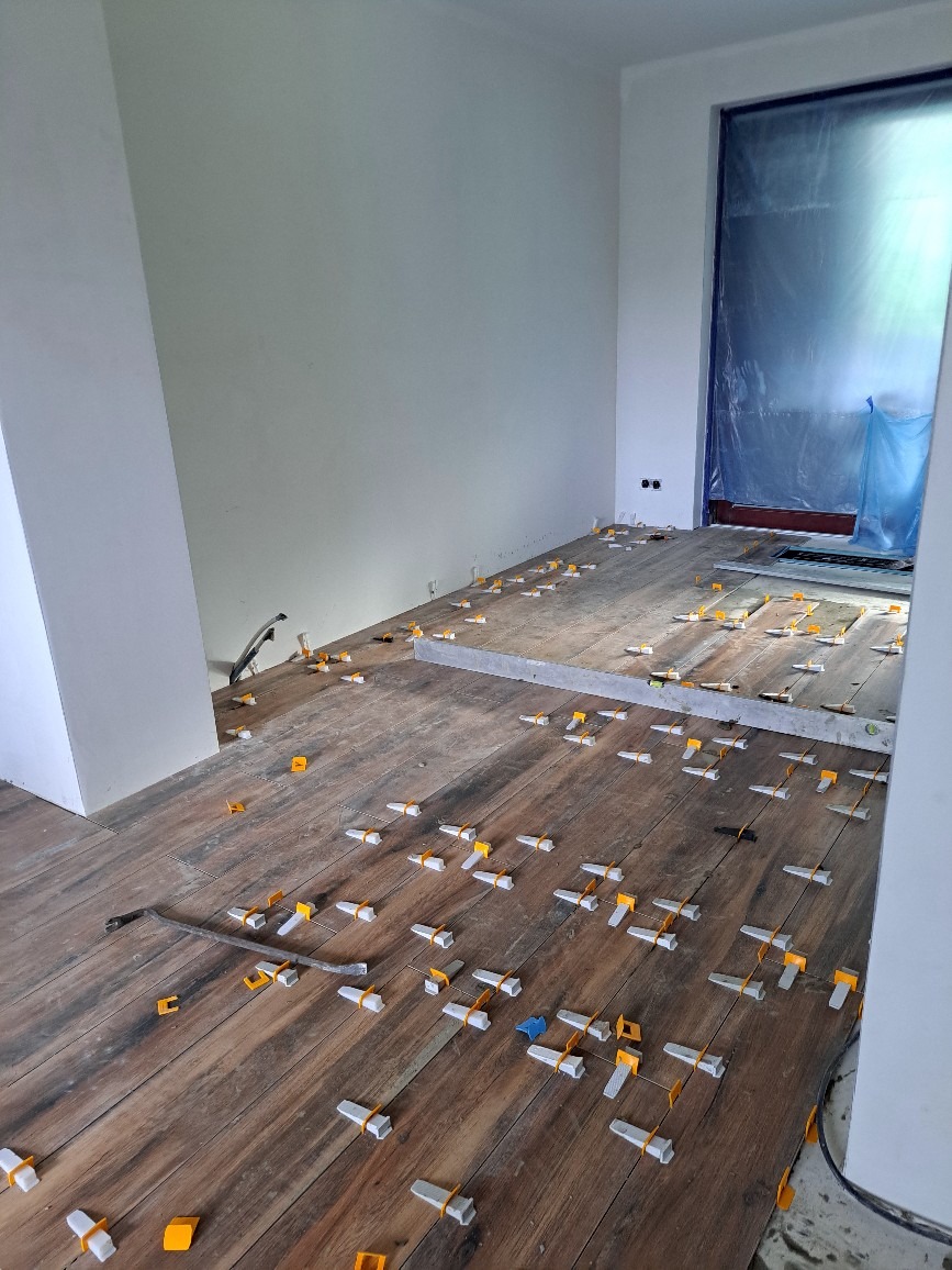 Układanie drewnopodobnych płytek podłogowych z użyciem systemu poziomowania, widoczna poziomica i narzędzia, zabezpieczone folią drzwi w tle.