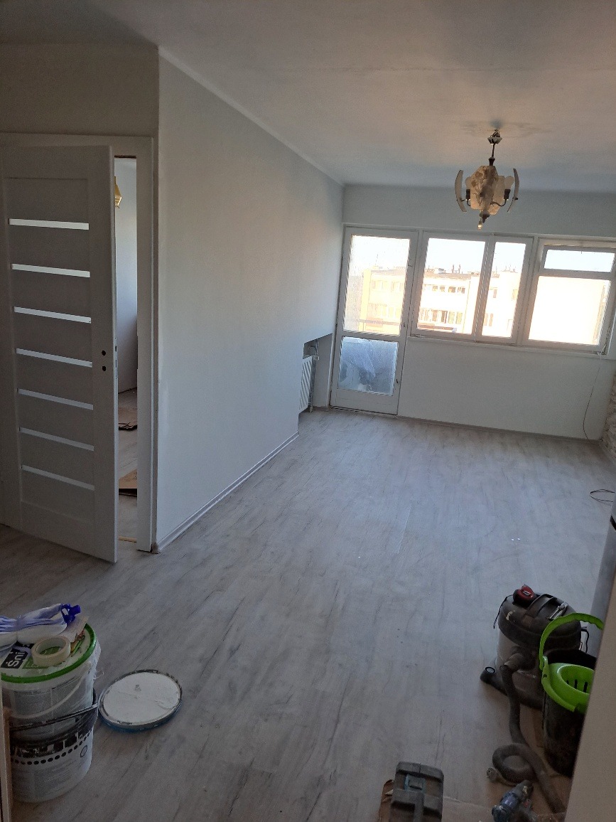 Wnętrze pokoju po malowaniu ścian na biało, widoczne otwarte drzwi z przeszkleniami, wiadro z farbą i akcesoria malarskie na podłodze, okno balkonowe w tle.