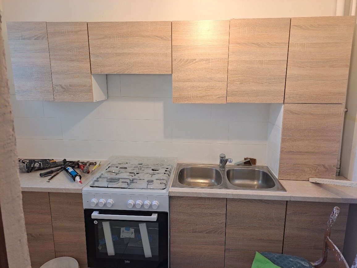 Niedokończona kuchnia z jasnymi drewnianymi szafkami, białą glazurą i nową kuchenką gazową z folią ochronną, widoczne narzędzia na blacie.