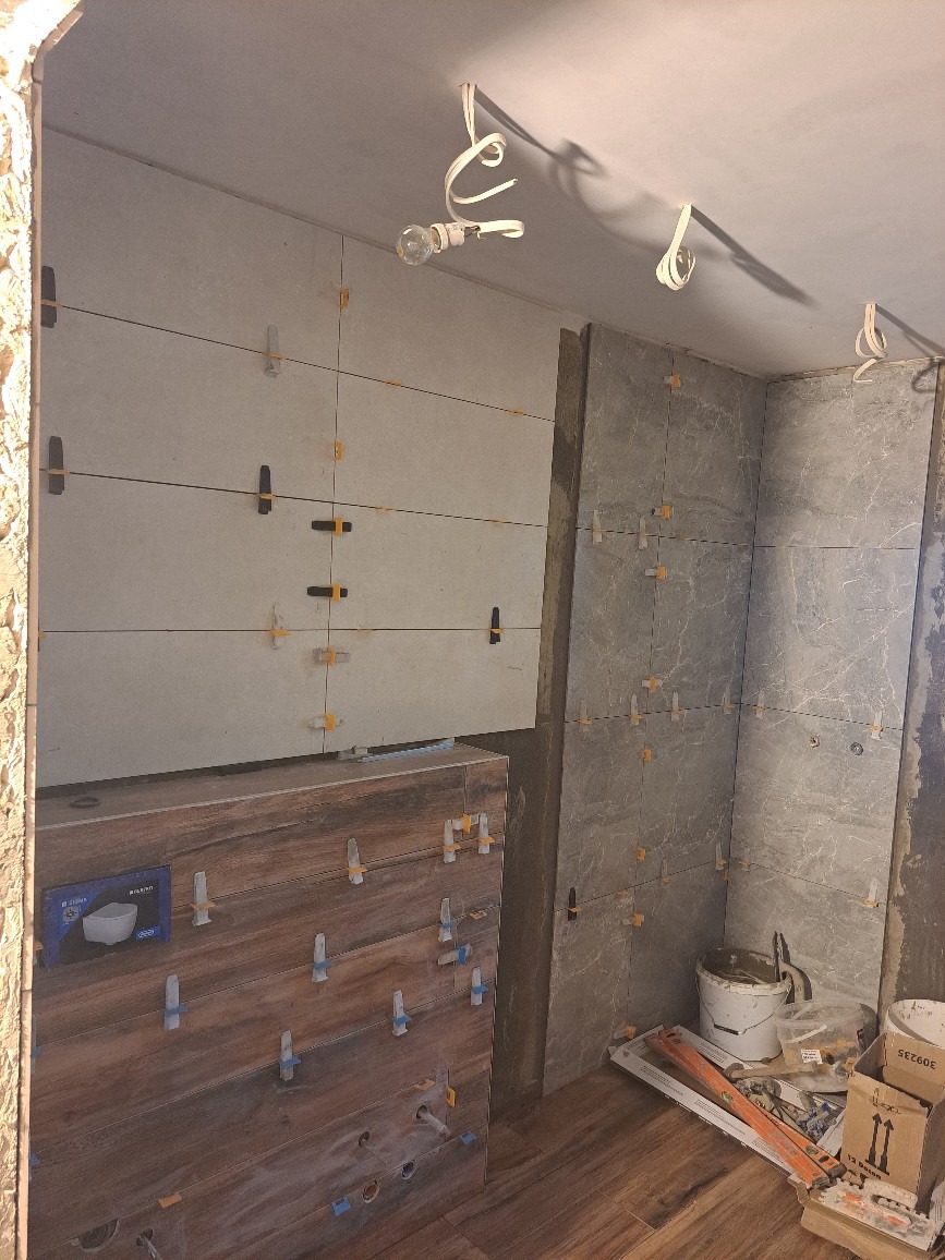 Świeżo ułożone, szare płytki na ścianach łazienki, zabezpieczone krzyżykami dystansowymi, z widocznymi przewodami oświetleniowymi na suficie i częściowo obudowaną szafką pod umywalkę z dekoracyjną...