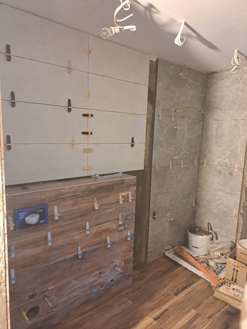 Proces układania płytek w łazience: szare płytki ścienne w trakcie montażu z użyciem krzyżyków dystansowych, drewnopodobna zabudowa pod umywalkę z wywierconymi otworami na instalację, widoczne...