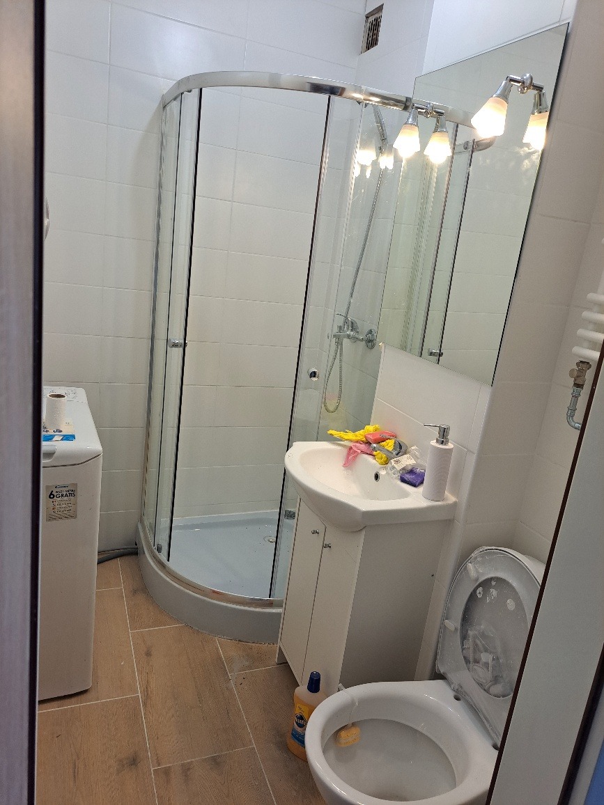 Niewielka łazienka z kabiną prysznicową, umywalką z szafką, pralką i toaletą w trakcie sprzątania, widoczny płyn do czyszczenia i gąbka.