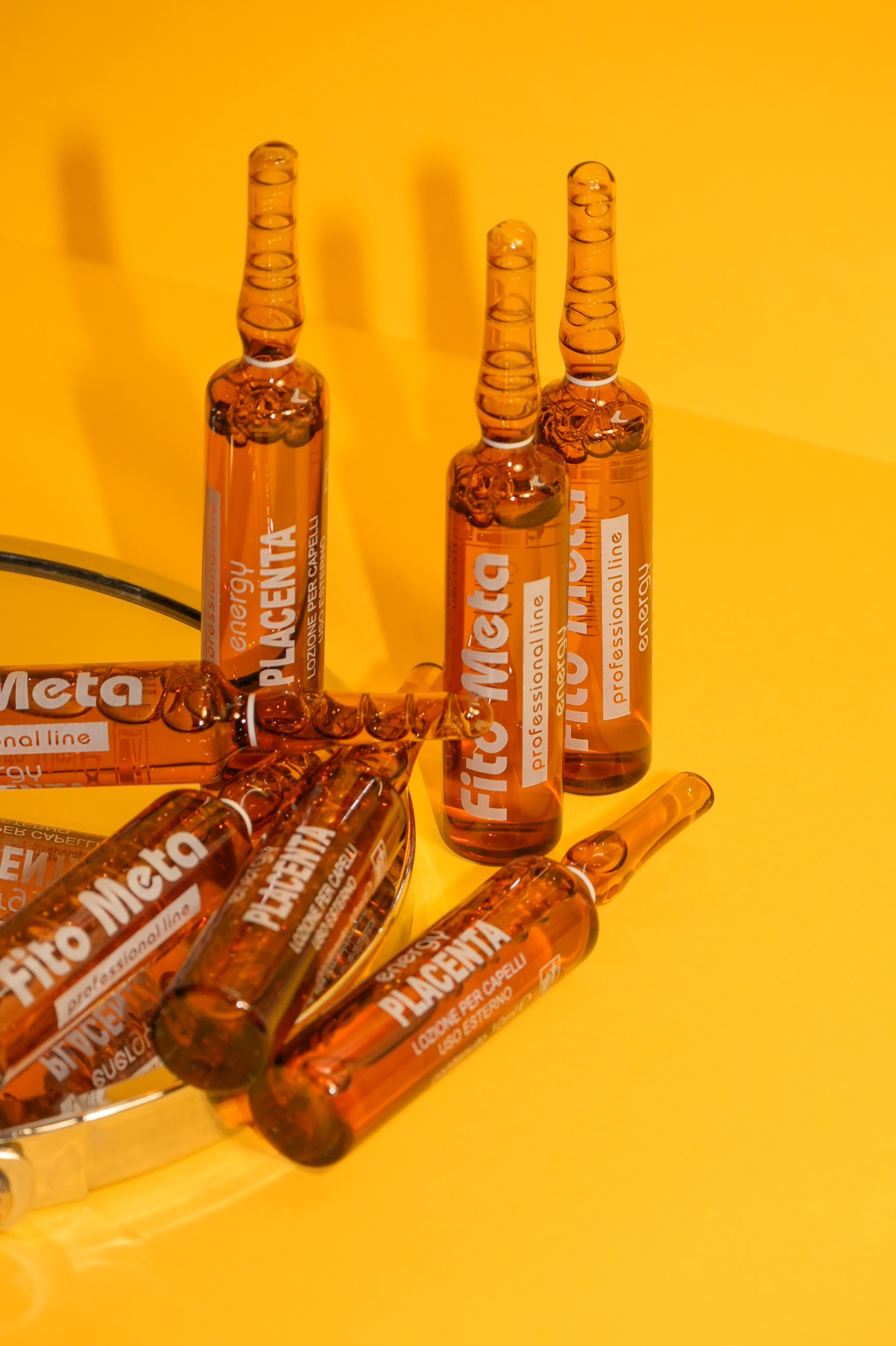 Szklane ampułki z płynem do włosów Fito Meta Placenta na żółtym tle, niektóre leżą na srebrnej tacce, ukazując etykiety z nazwą i przeznaczeniem produktu.