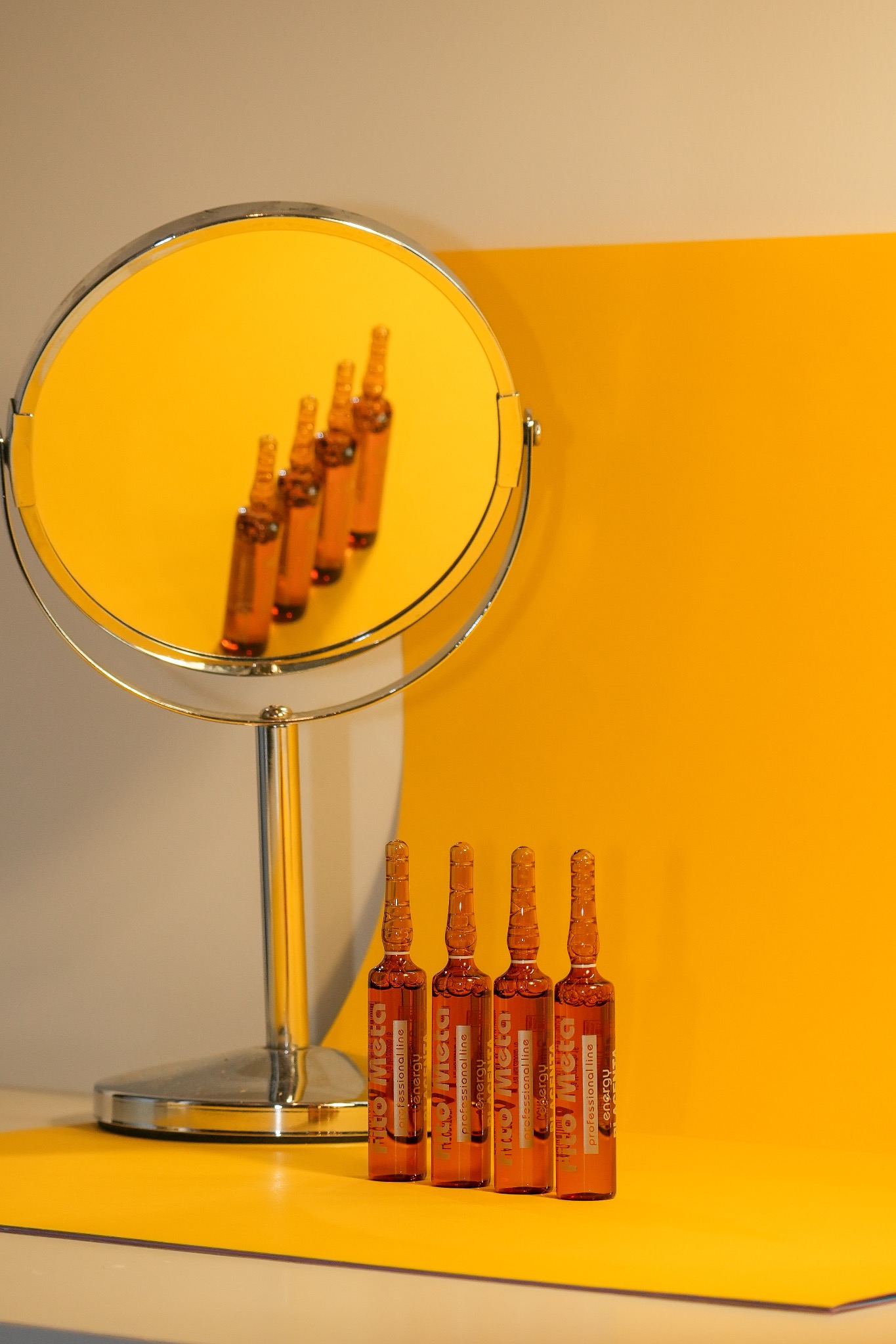 Cztery ampułki z płynem stojące na żółto-białym tle, odbicie w okrągłym lustrze ukazuje ich rząd w oddali.