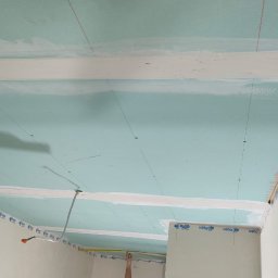 Remdom - Mężczyzna mierzy odległość taśmą mierniczą na suficie z zamontowaną płytą kartonowo-gipsową, widoczne linie pomocnicze i taśmy Rigips