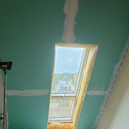 Remdom - Montaż płyty kartonowo-gipsowej na poddaszu z oknem dachowym, widoczny szpachlowany łączenie płyt, drabina z wkrętarką, wełna mineralna pod oknem