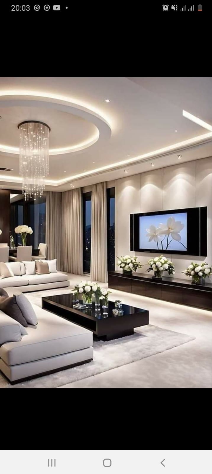 Elegancki salon z podwieszanym sufitem z oświetleniem LED, kryształowym żyrandolem, beżowymi zasłonami i meblami w odcieniach bieli i czerni.