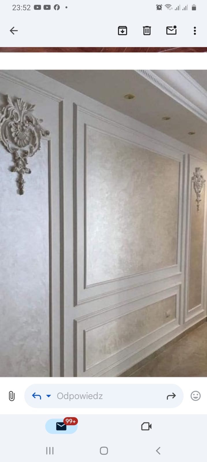 Elegancka ściana z dekoracyjnymi sztukateriami i panelami w odcieniach bieli i beżu, oświetlona punktowymi lampami sufitowymi.