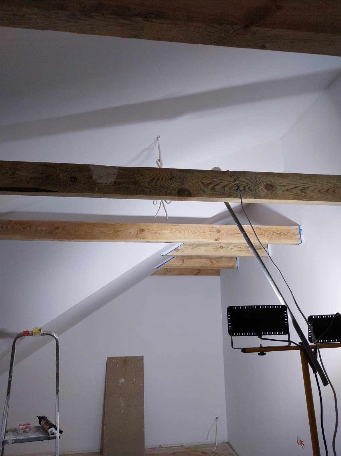 Wnętrze poddasza w trakcie prac wykończeniowych: widoczne drewniane belki stropowe, oświetlenie budowlane, drabina i płyta karton-gips oparta o ścianę, ściany wstępnie przygotowane pod dalsze prace.