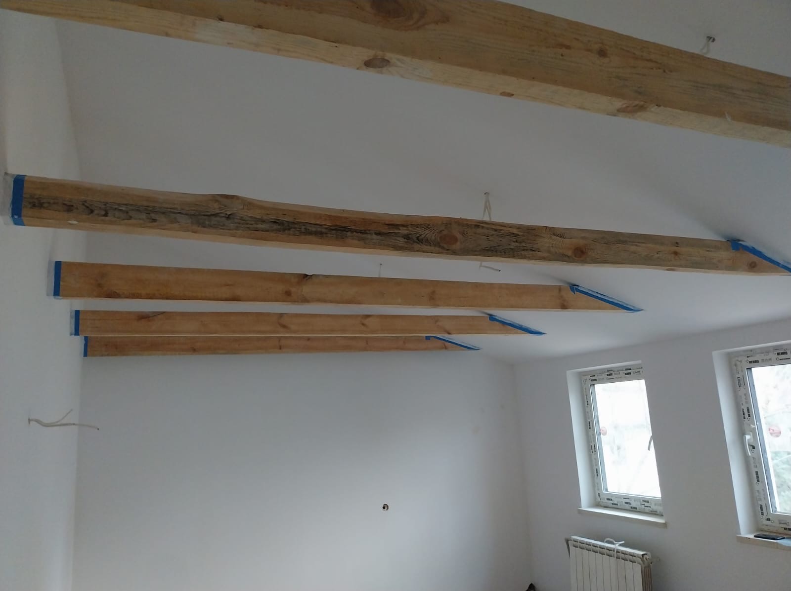 Montaż drewnianych belek stropowych, zabezpieczonych niebieską taśmą malarską, na białym suficie w jasnym pomieszczeniu z dwoma oknami i grzejnikiem.