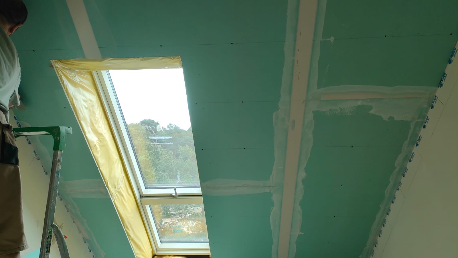Montaż płyt kartonowo-gipsowych na poddaszu z oknem dachowym, widoczne łączenia płyt wzmocnione taśmą i częściowo zaszpachlowane, okno zabezpieczone folią malarską, pracownik na drabinie.
