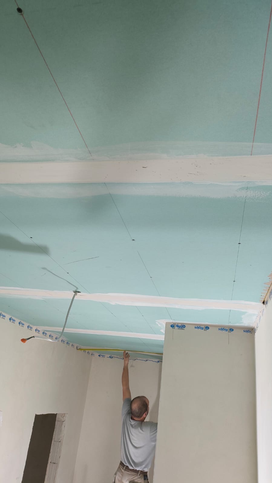 Mężczyzna mierzy odległość taśmą mierniczą na suficie z zamontowaną płytą kartonowo-gipsową, widoczne linie pomocnicze i taśmy Rigips