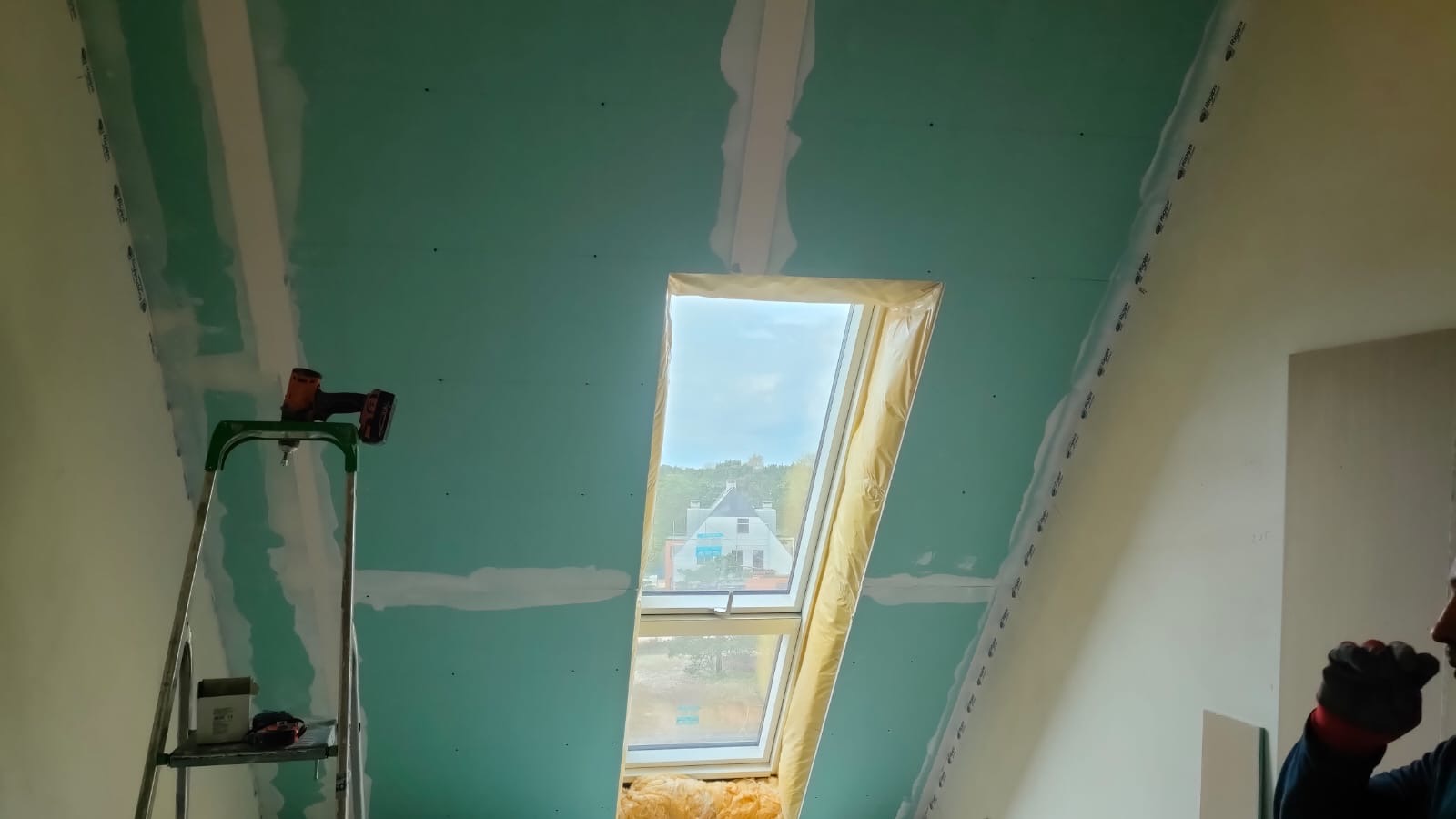 Montaż płyty kartonowo-gipsowej na poddaszu z oknem dachowym, widoczny szpachlowany łączenie płyt, drabina z wkrętarką, wełna mineralna pod oknem