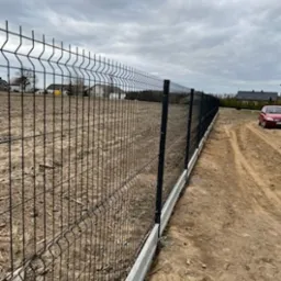 Nowoczesne ogrodzenie panelowe z podmurówką betonową, widok z perspektywy gruntu, na tle pochmurnego nieba i zabudowań w oddali.