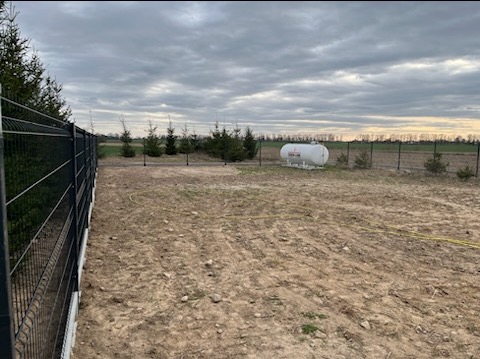 Metalowe ogrodzenie panelowe na nierównym terenie z widocznym zbiornikiem gazu i młodymi drzewkami iglastymi w tle, pochmurne niebo.