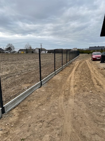 Nowoczesne ogrodzenie panelowe z podmurówką betonową, widoczne ślady opon na ziemi, w tle zabudowania wiejskie pod pochmurnym niebem.