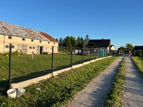 Nowe ogrodzenie panelowe z podmurówką betonową w wiejskiej scenerii. Widoczny budynek gospodarczy w tle i osoba w oddali. Słoneczny dzień.