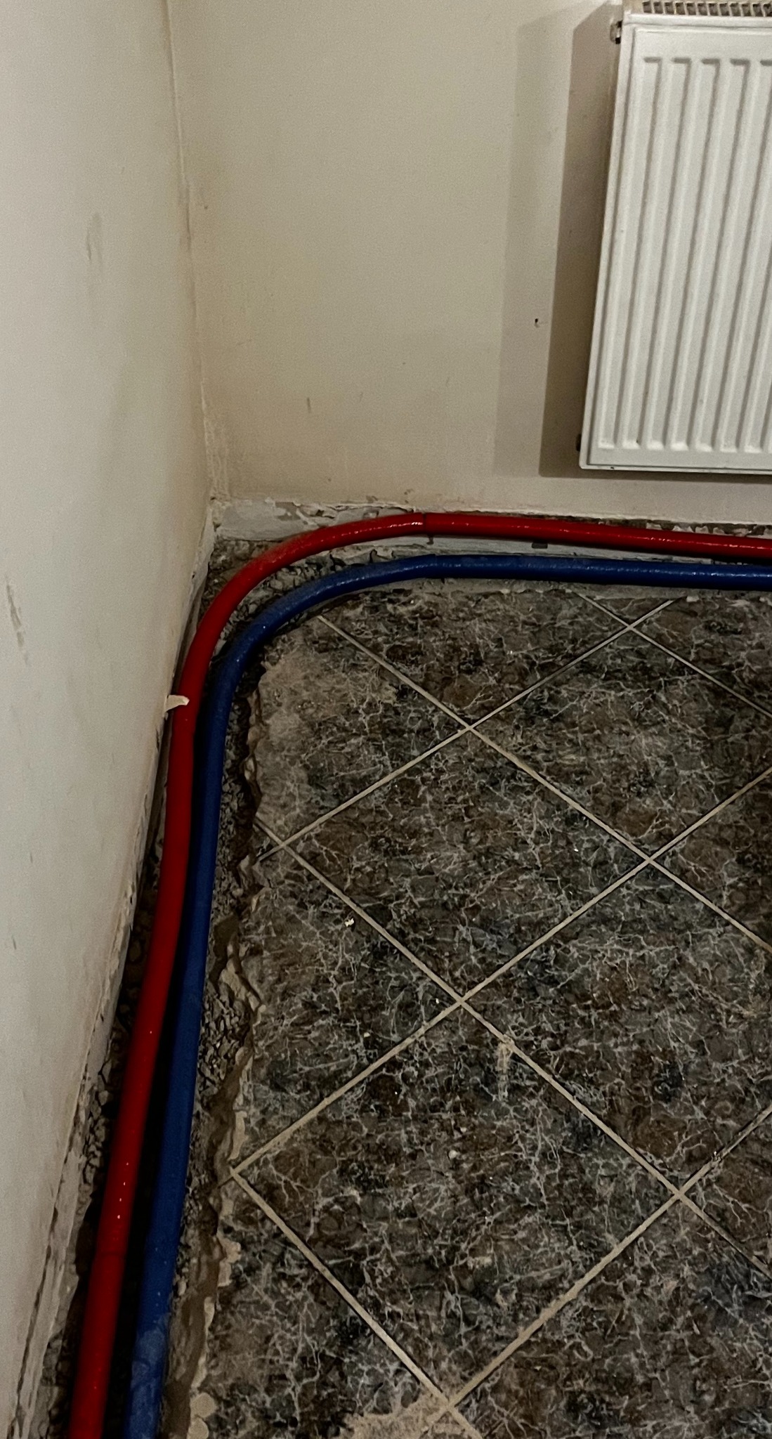 Czerwona i niebieska rura instalacji grzewczej ułożona wzdłuż ściany na podłodze z płytek podczas prac w Olsztynie, widoczny grzejnik.