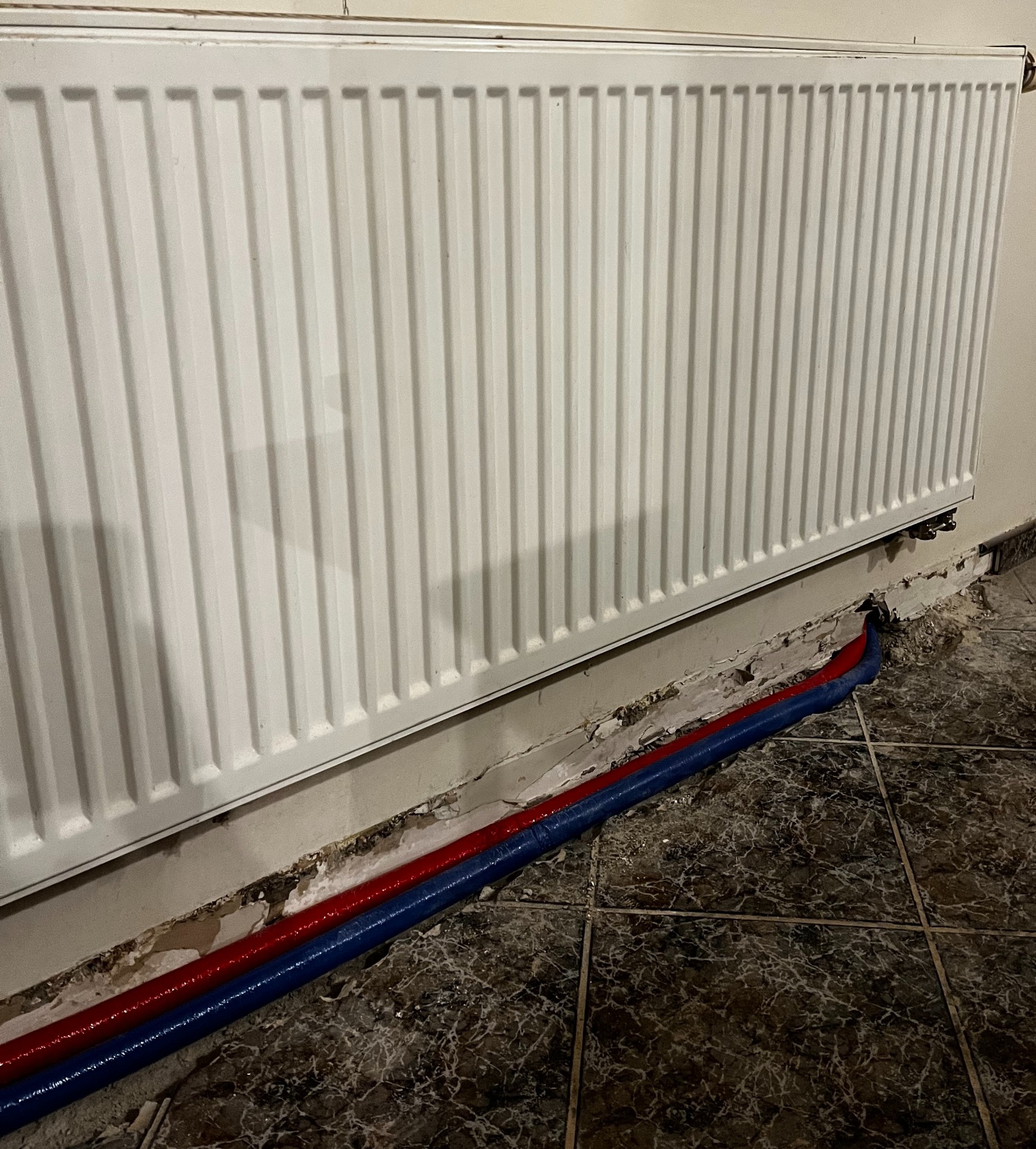 Biały grzejnik panelowy z podłączonymi rurami w kolorze niebieskim i czerwonym, widoczne ślady po kuciu w ścianie, marmurowe płytki podłogowe.