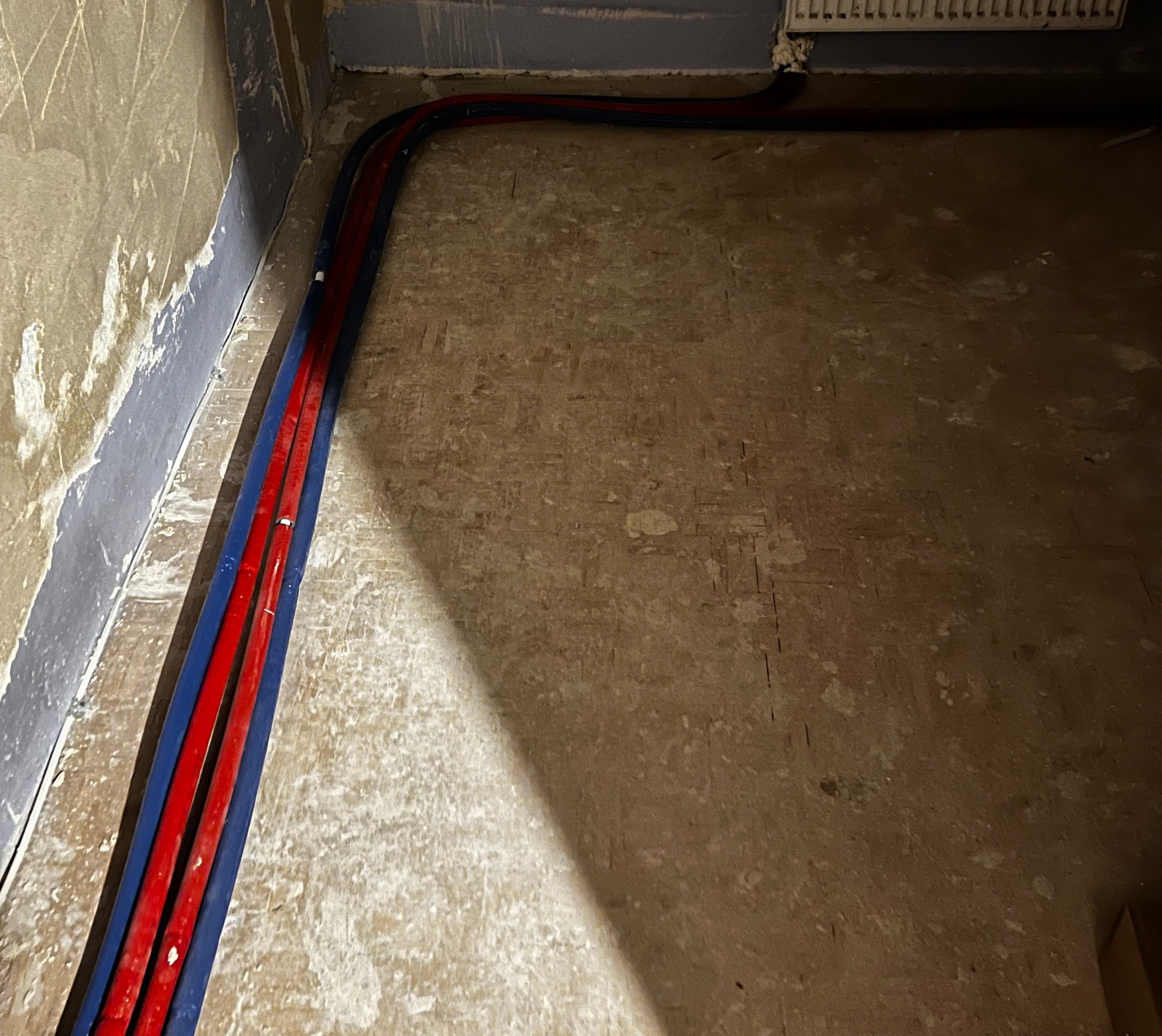 Wewnętrzne rury grzewcze w kolorze czerwonym i niebieskim ułożone wzdłuż ściany w surowym stanie, przed wykończeniem podłogi i ścian.