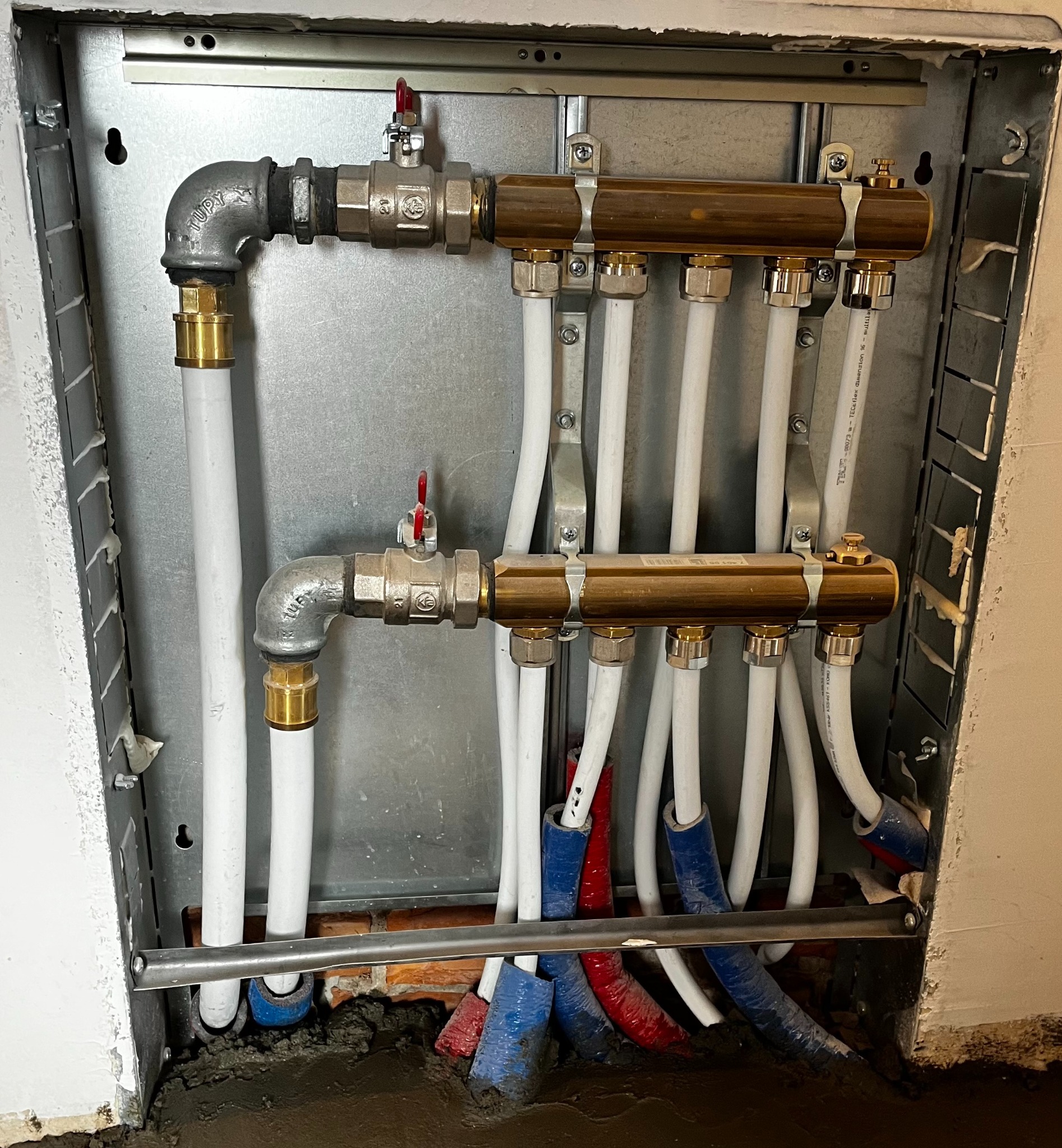 Rozdzielacz ogrzewania podłogowego w metalowej szafce, widoczne rury PEX w izolacji niebieskiej i czerwonej, przyłącza mosiężne i zawory kulowe z czerwonymi rączkami.