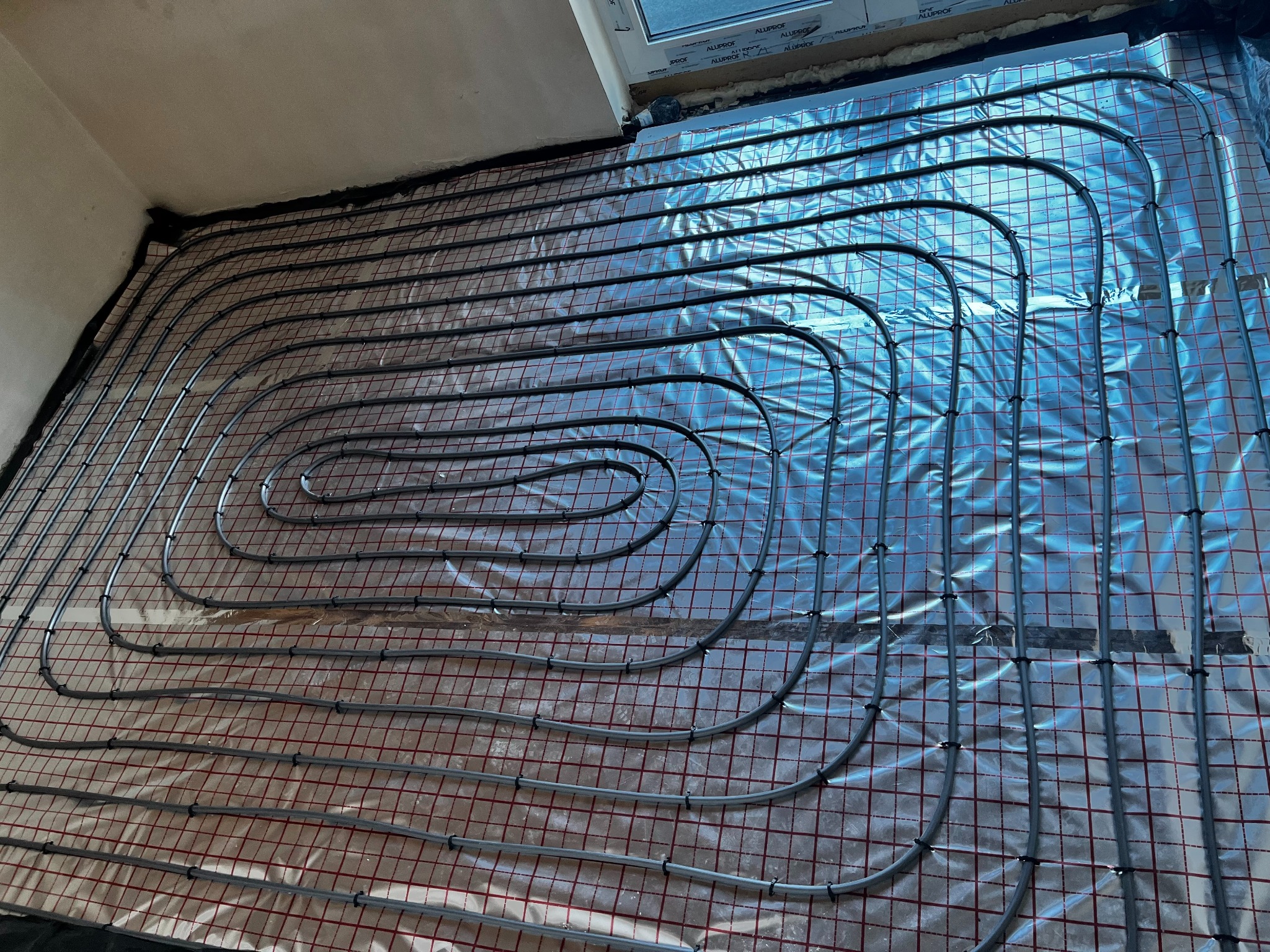 Szare rury ogrzewania podłogowego ułożone spiralnie na folii izolacyjnej z czerwoną siatką, widok z góry w trakcie montażu w domu w Barwinach o powierzchni 260 m2.