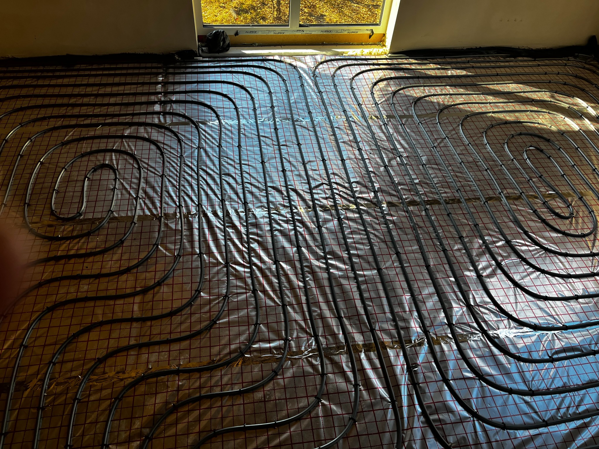 Instalacja ogrzewania podłogowego z rurkami ułożonymi spiralnie na folii refleksyjnej z widocznym oknem w tle.