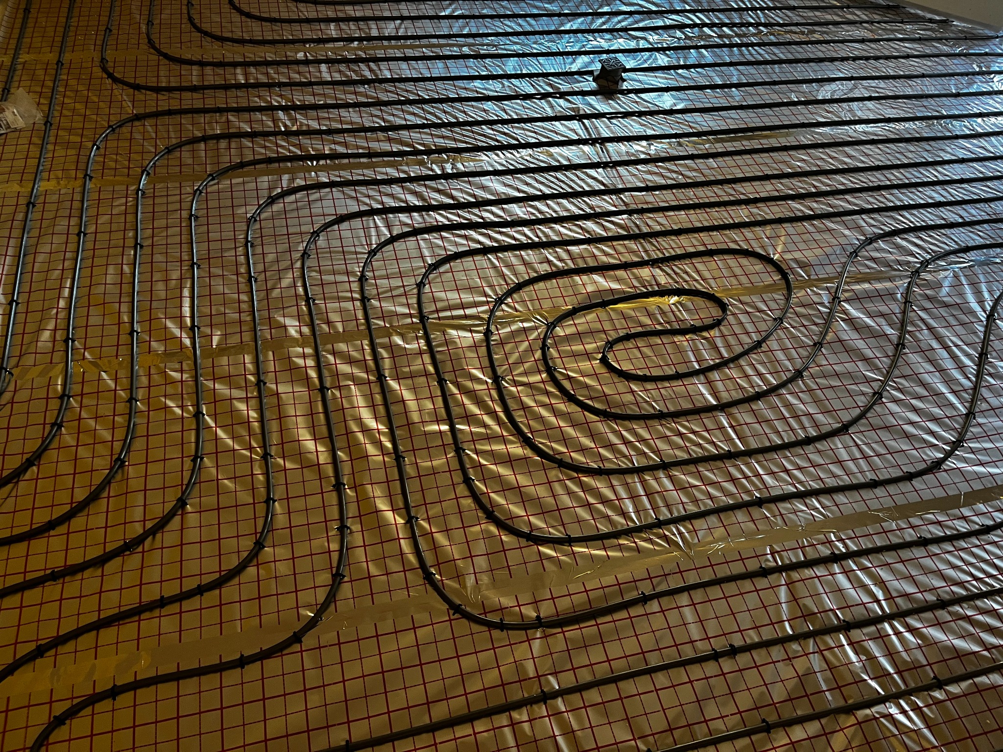 Rury ogrzewania podłogowego ułożone na folii z siatką, widoczne elementy mocujące, odbicie światła na folii.