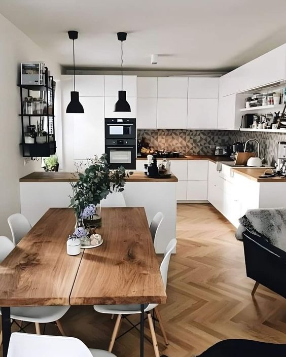 Jasna kuchnia z białymi szafkami, drewnianym blatem i geometryczną mozaiką na ścianie, uzupełniona drewnianym stołem z kwiatami i czarnymi lampami wiszącymi.