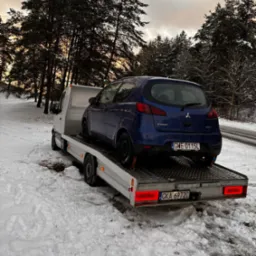 Niebieski samoch&oacute;d Mitsubishi Colt zabezpieczony pasami na platformie lawety w zimowej scenerii, widok z tyłu, tablice rejestracyjne GWE 0115L, śnieg na drodze i drzewach.