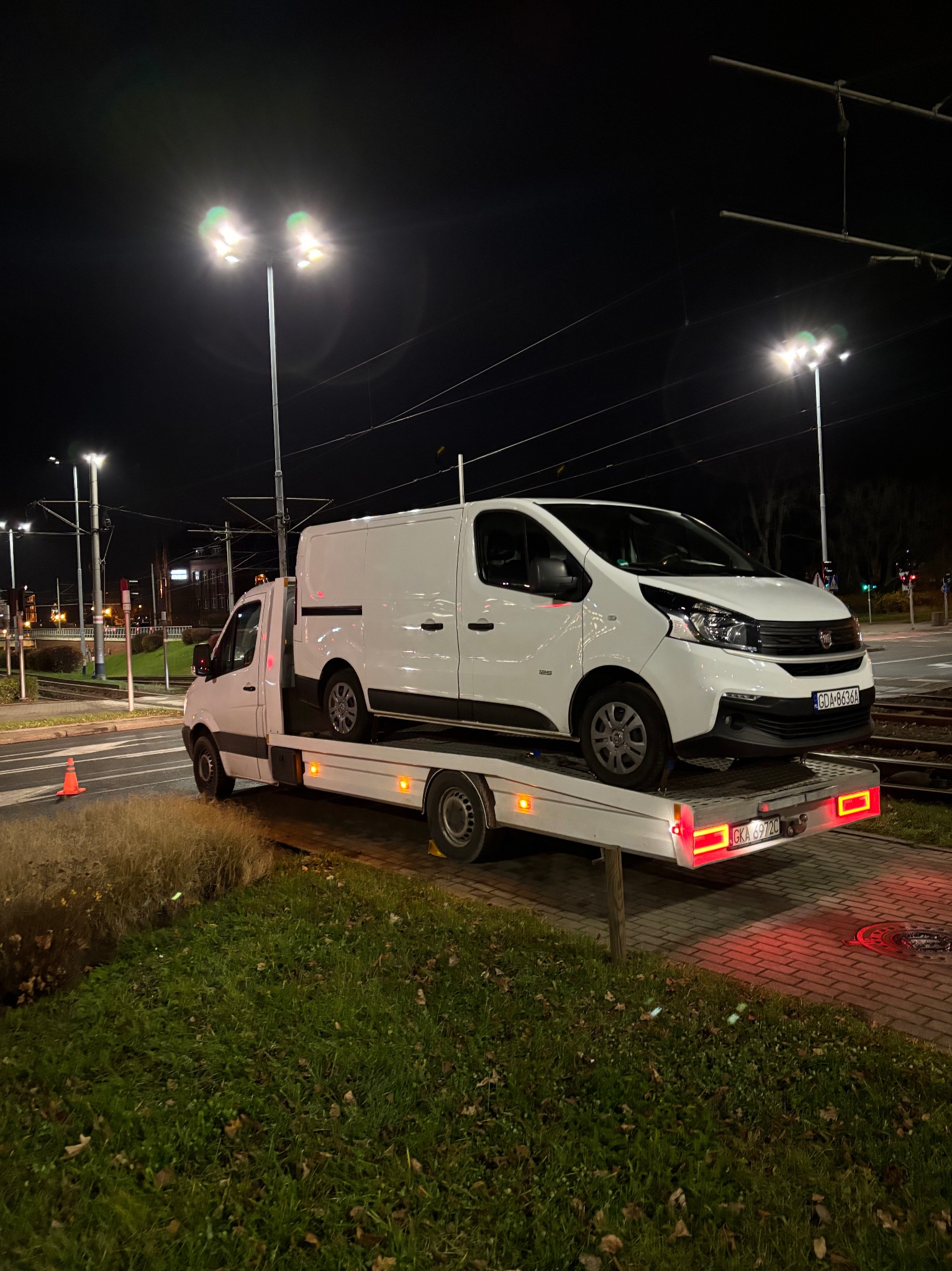 Biały bus transportowany na lawecie na tle nocnego miasta z torami tramwajowymi.