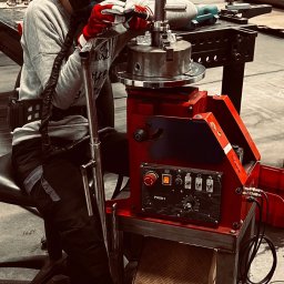 L-WELD Krystian Lenzer - Operator w masce spawalniczej, w czerwonych rękawicach, pracuje przy czerwono-czarnym urządzeniu z panelem sterowania, obrabiając metalowy element w warsztacie.