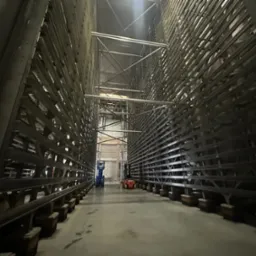 Wnętrze magazynu z regałami metalowymi, widok na wózek widłowy i podnośnik koszowy w oddali.