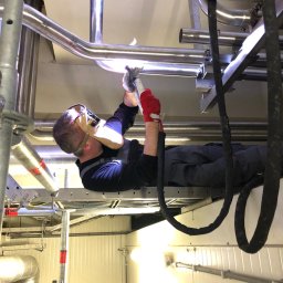 L-WELD Krystian Lenzer - Pracownik w masce spawalniczej, leżąc na rusztowaniu, łączy rury ze stali nierdzewnej wewnątrz budynku przemysłowego, używając metody spawania TIG.