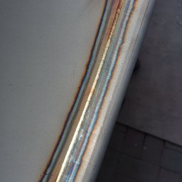 L-WELD Krystian Lenzer - Szlifowana powierzchnia metalowa z widocznym spawem o opalizujących barwach termicznych, położona na szarym tle.