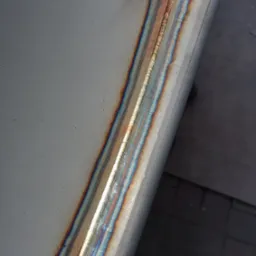 Szlifowana powierzchnia metalowa z widocznym spawem o opalizujących barwach termicznych, położona na szarym tle.