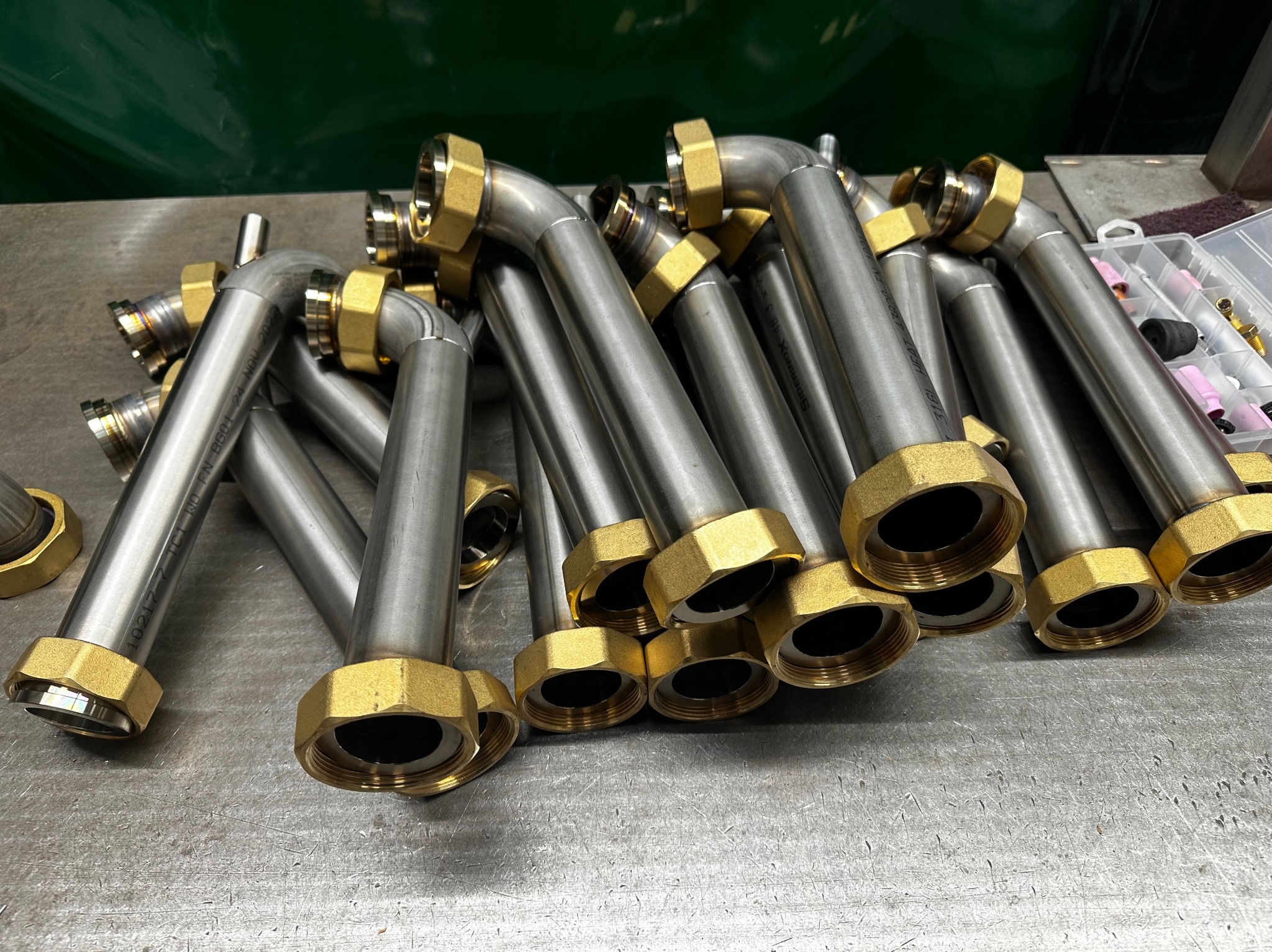 Srebrne metalowe rury z mosiężnymi nakrętkami ułożone na metalowym stole warsztatowym, widoczne narzędzia i akcesoria spawalnicze w tle.