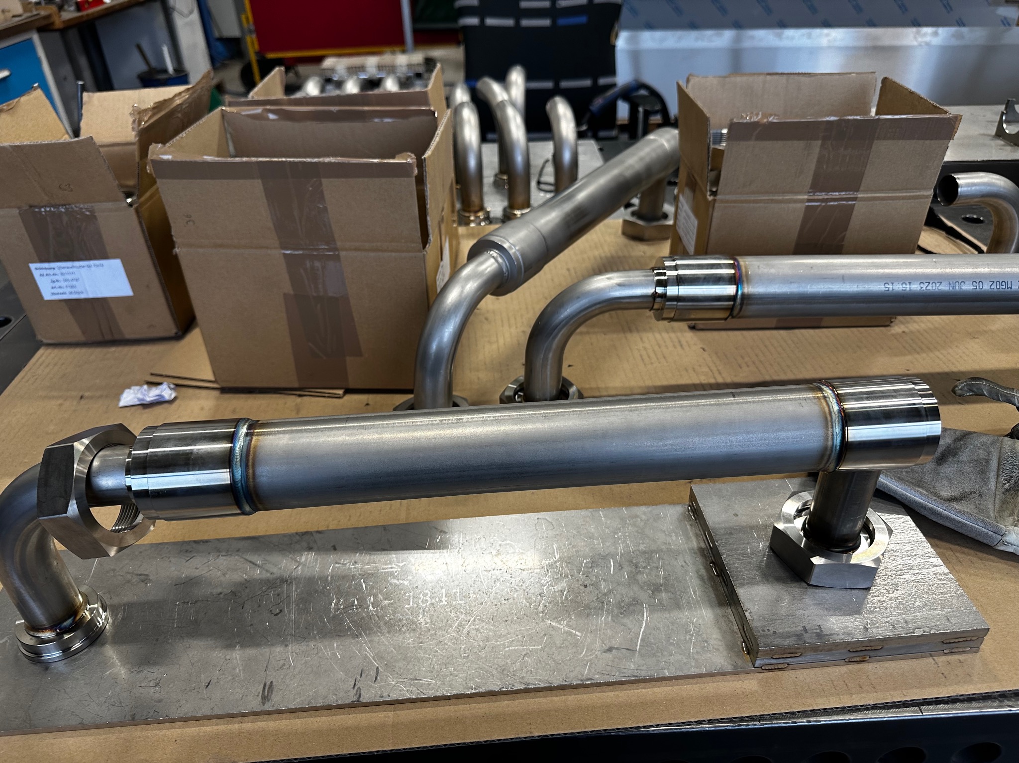 Spawane rury ze stali nierdzewnej z elementami gwintowanymi, ułożone na metalowej płycie w warsztacie, widoczne kartony i inne elementy rurowe w tle.