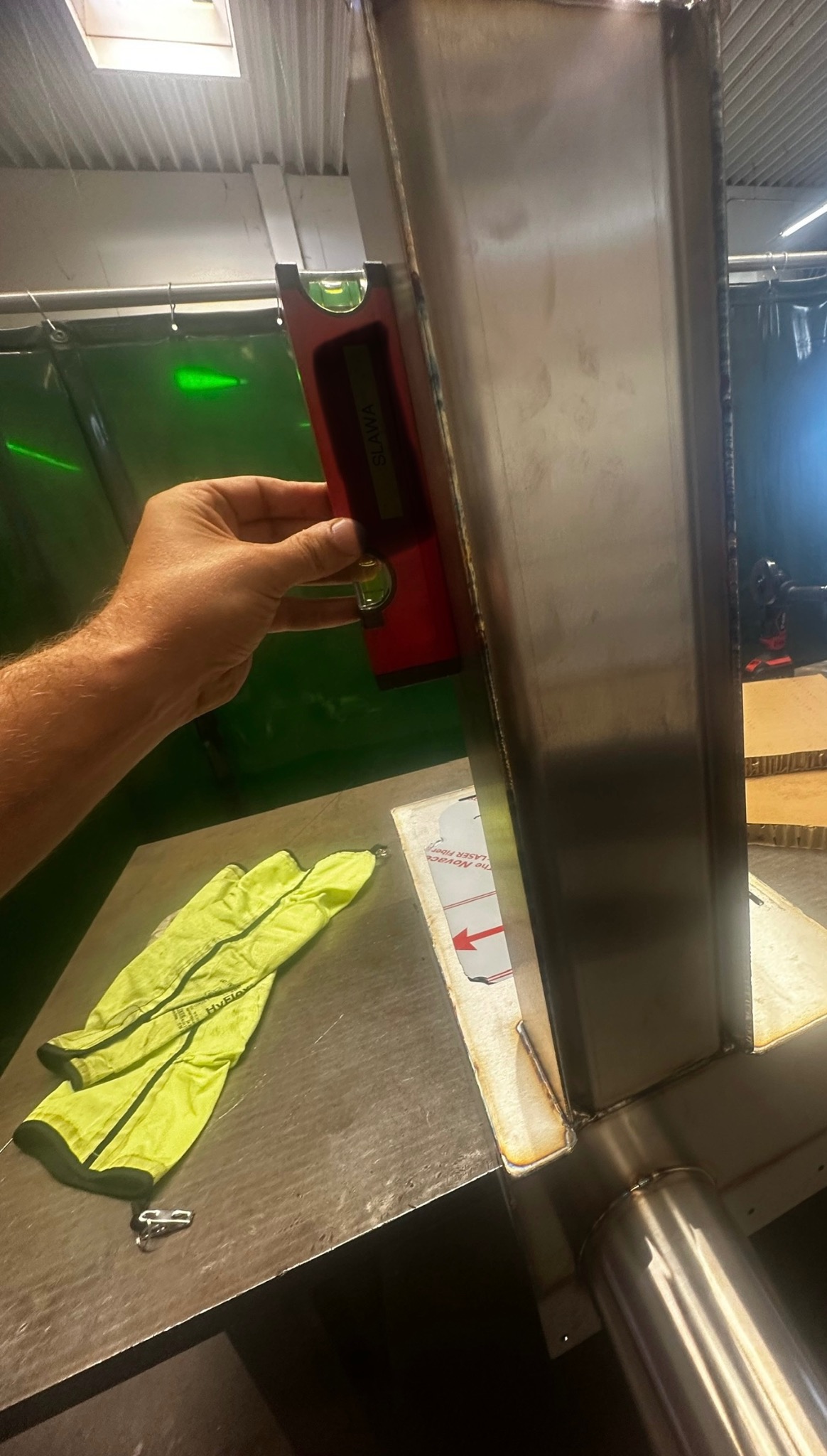 Sprawdzanie poziomu pionowego słupa ze stali nierdzewnej przy użyciu poziomicy w warsztacie, widoczne żółte rękawice robocze i osłona spawalnicza w tle.