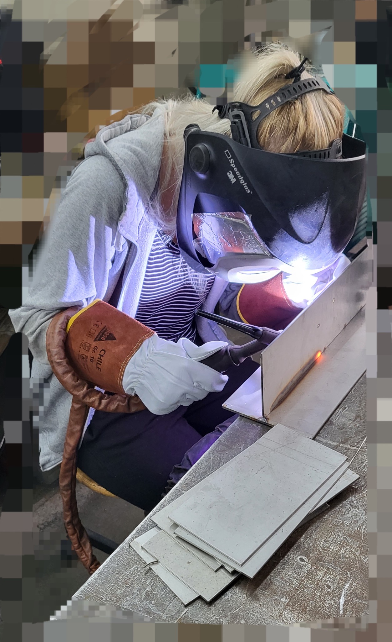 Kobieta w masce spawalniczej 3M Speedglas 9100XXi, rękawicach ochronnych i szarej bluzie z kapturem, wykonuje spawanie elementu metalowego w warsztacie.