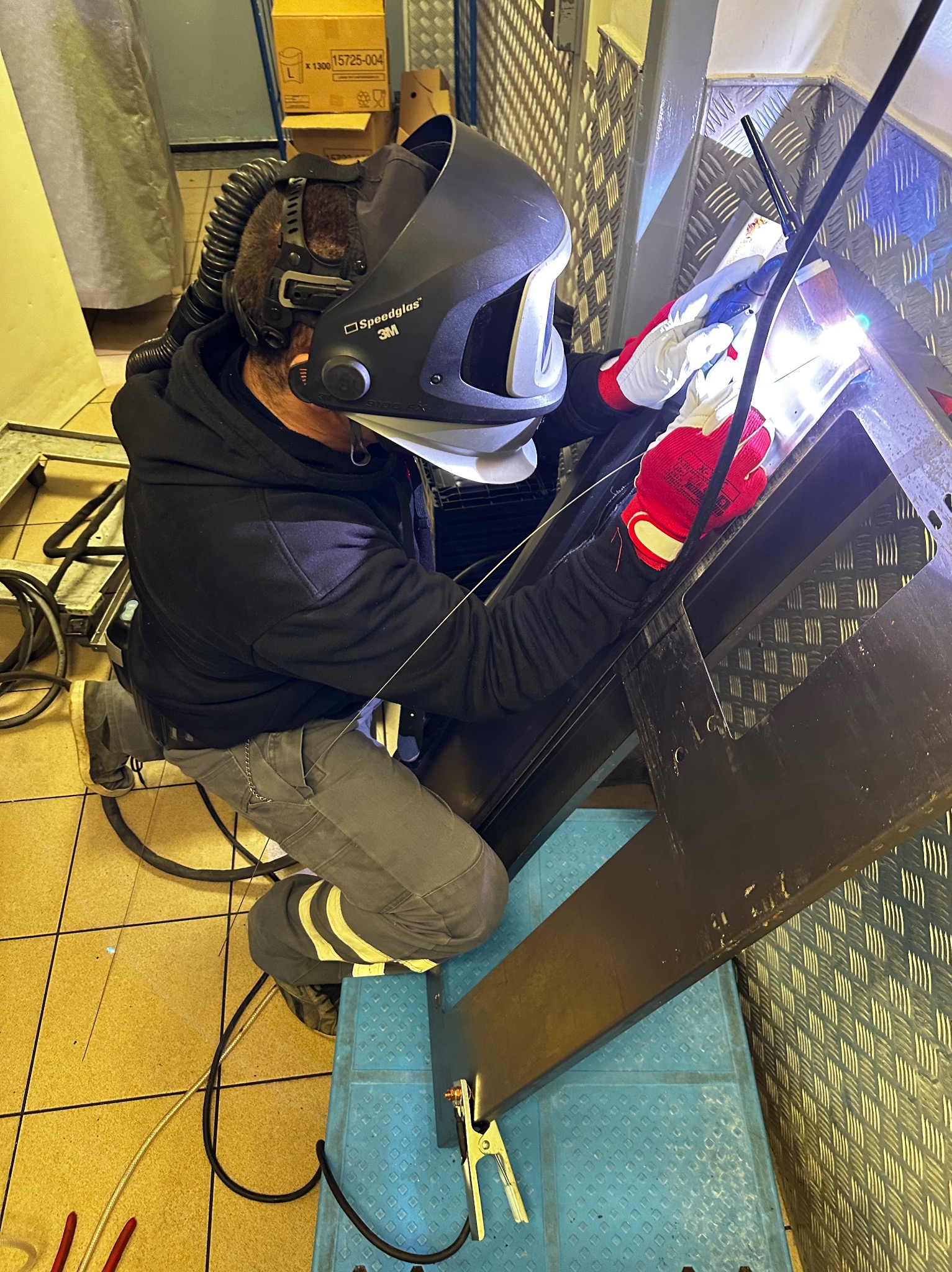 Pracownik w masce spawalniczej 3M Speedglas, w czerwonych rękawicach ochronnych, spawa metalową konstrukcję schodów na tle blachy ryflowanej i żółtej podłogi.