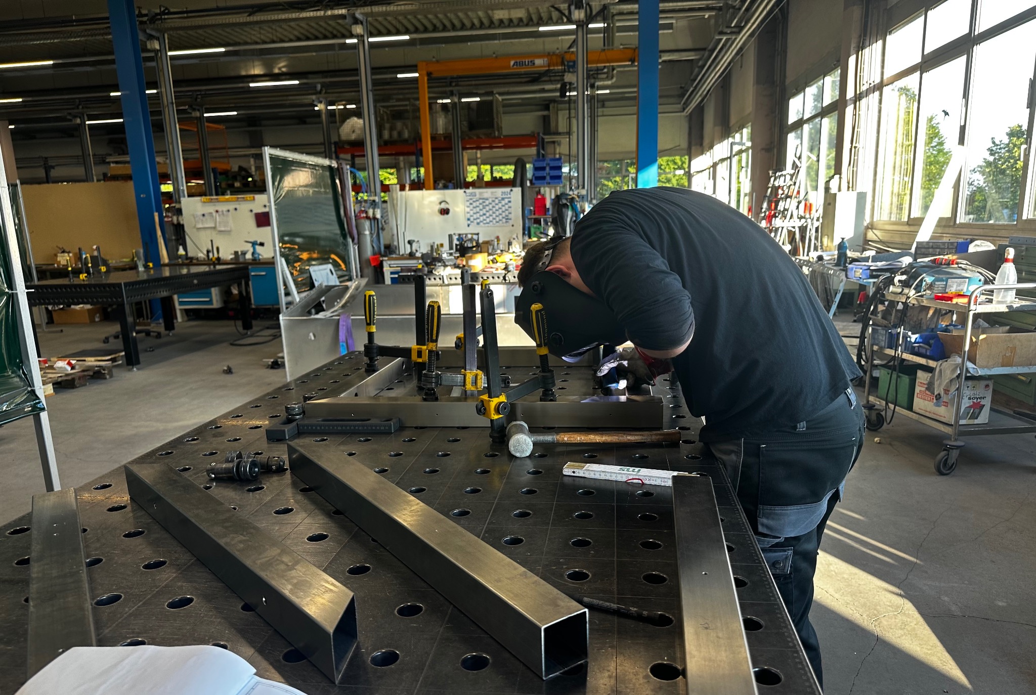 Pracownik w masce spawalniczej łączy metalowe profile na perforowanym stole warsztatowym przy użyciu klem i narzędzi pomiarowych w hali produkcyjnej.