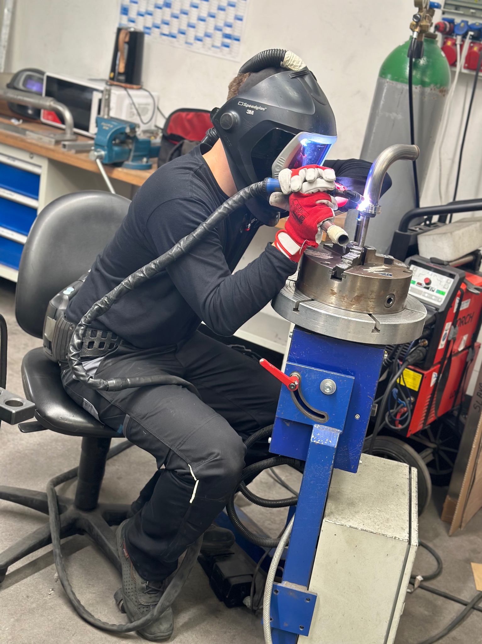 Pracownik w masce spawalniczej 3M, w czerwonych rękawicach, wykonuje precyzyjne łączenie rur na specjalistycznym stanowisku z obrotowym uchwytem, w tle butla z gazem i spawarka.