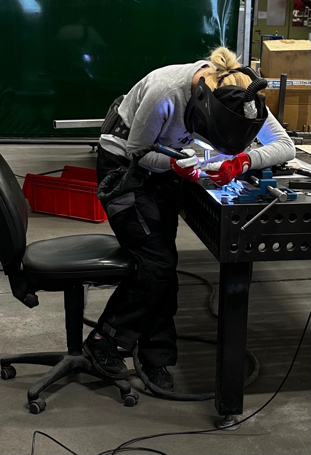 Spawaczka w masce ochronnej, z czerwonymi rękawicami, spawająca metalowy element na perforowanym stole spawalniczym w warsztacie.