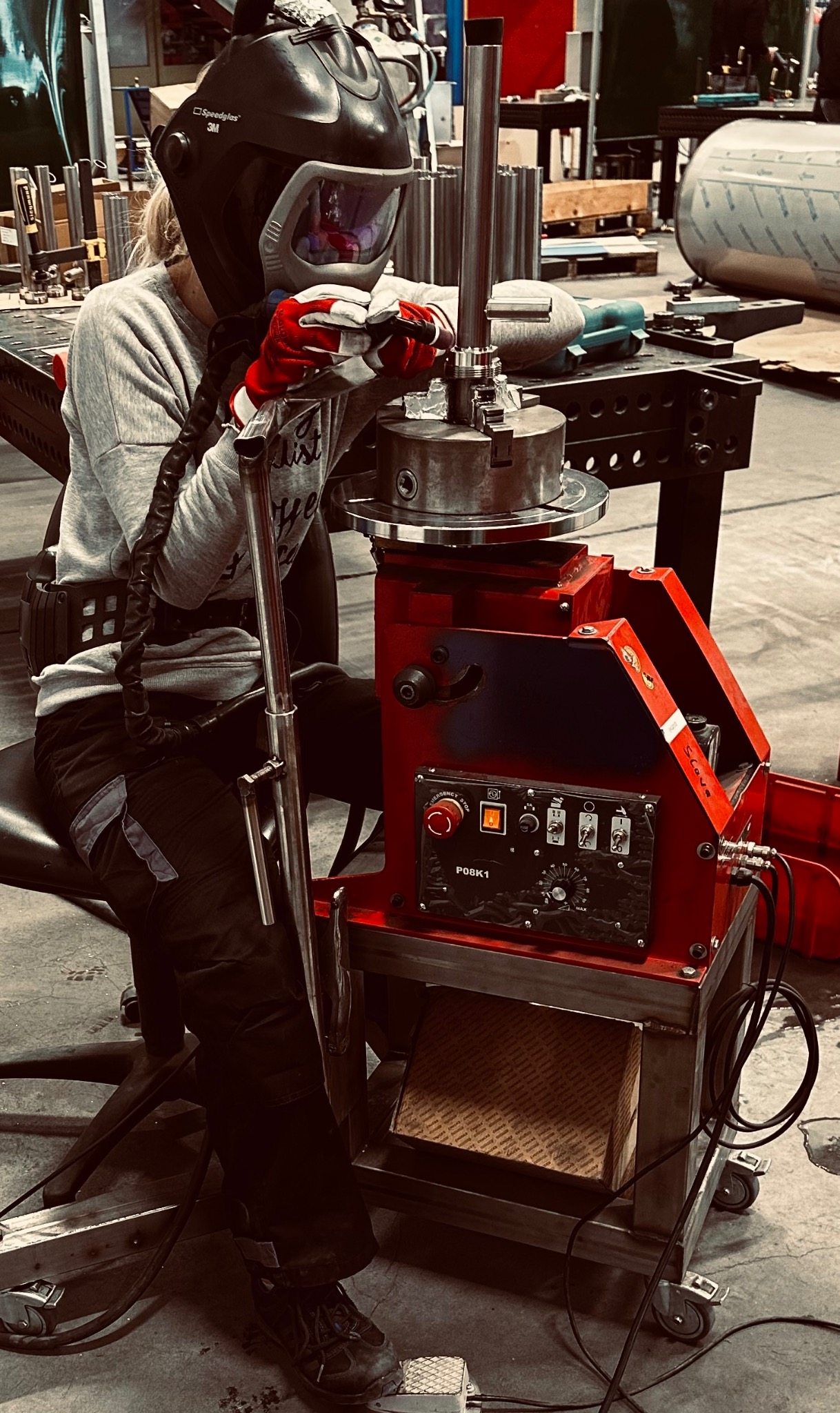 Operator w masce spawalniczej, w czerwonych rękawicach, pracuje przy czerwono-czarnym urządzeniu z panelem sterowania, obrabiając metalowy element w warsztacie.