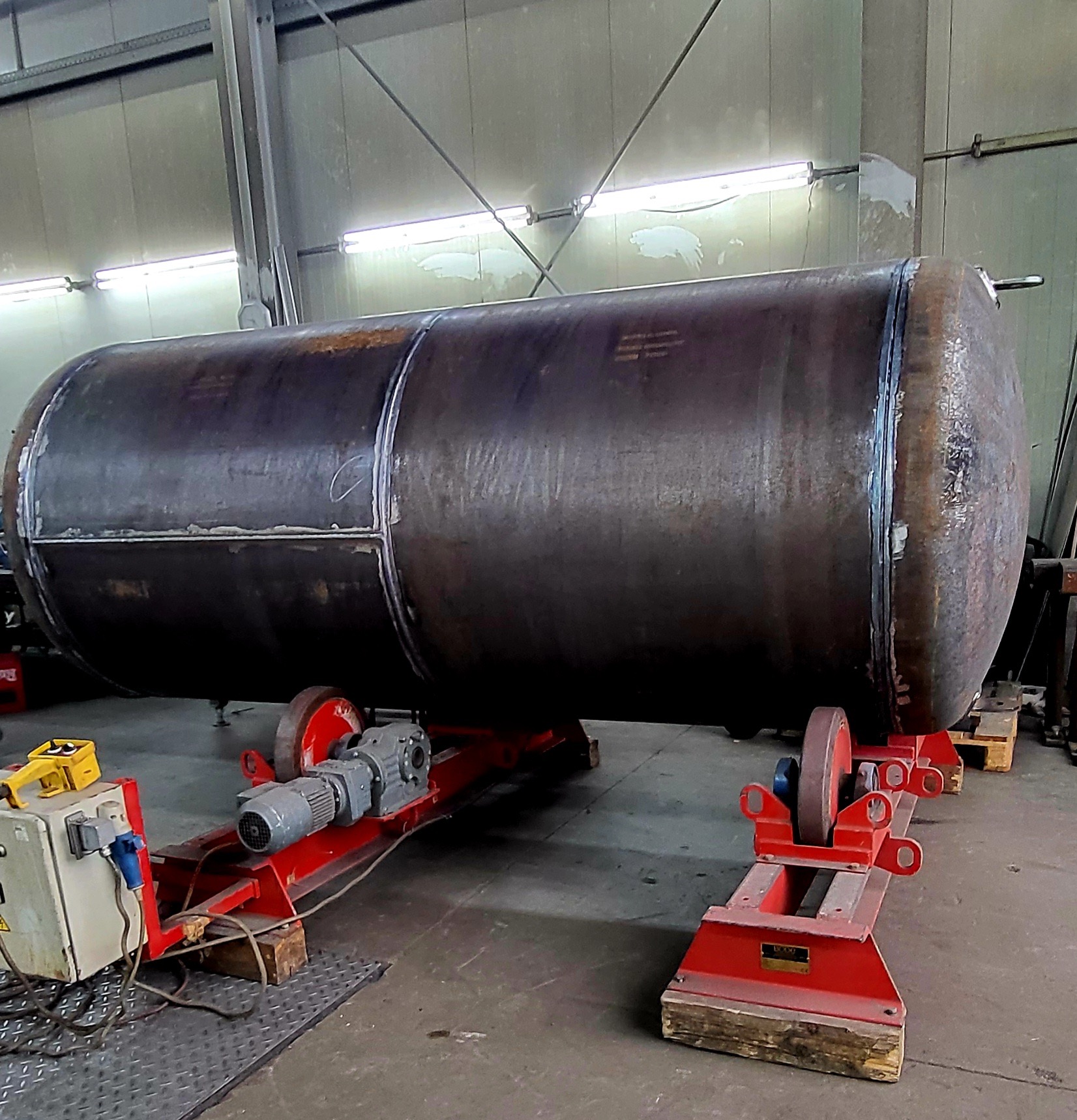 Cylindryczny, metalowy zbiornik na czerwonym stanowisku rolkowym w hali produkcyjnej, widoczne ślady spawania na łączeniach segmentów.