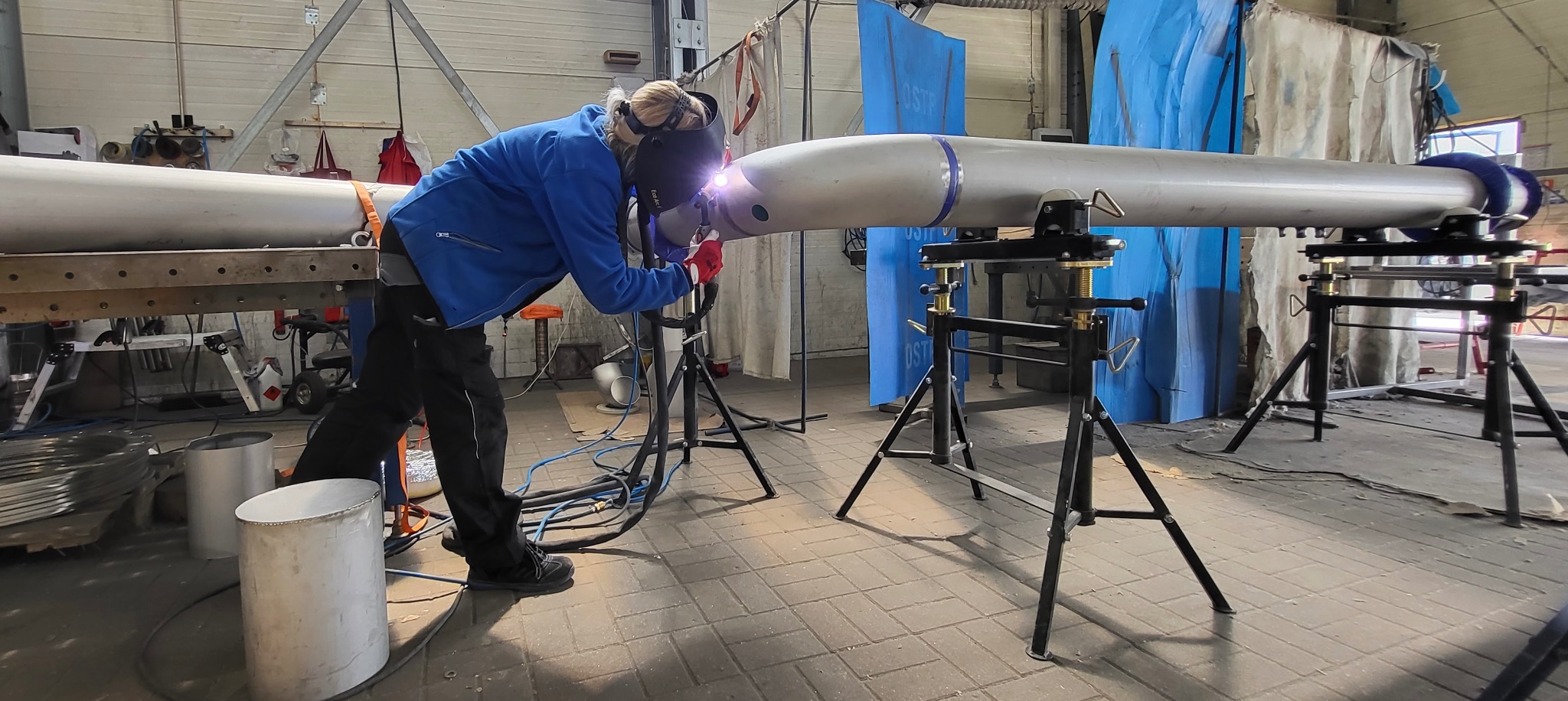 Pracownik w masce spawalniczej łączy dwa metalowe elementy o cylindrycznym kształcie za pomocą spawarki, elementy te są zamocowane na specjalnych stojakach.