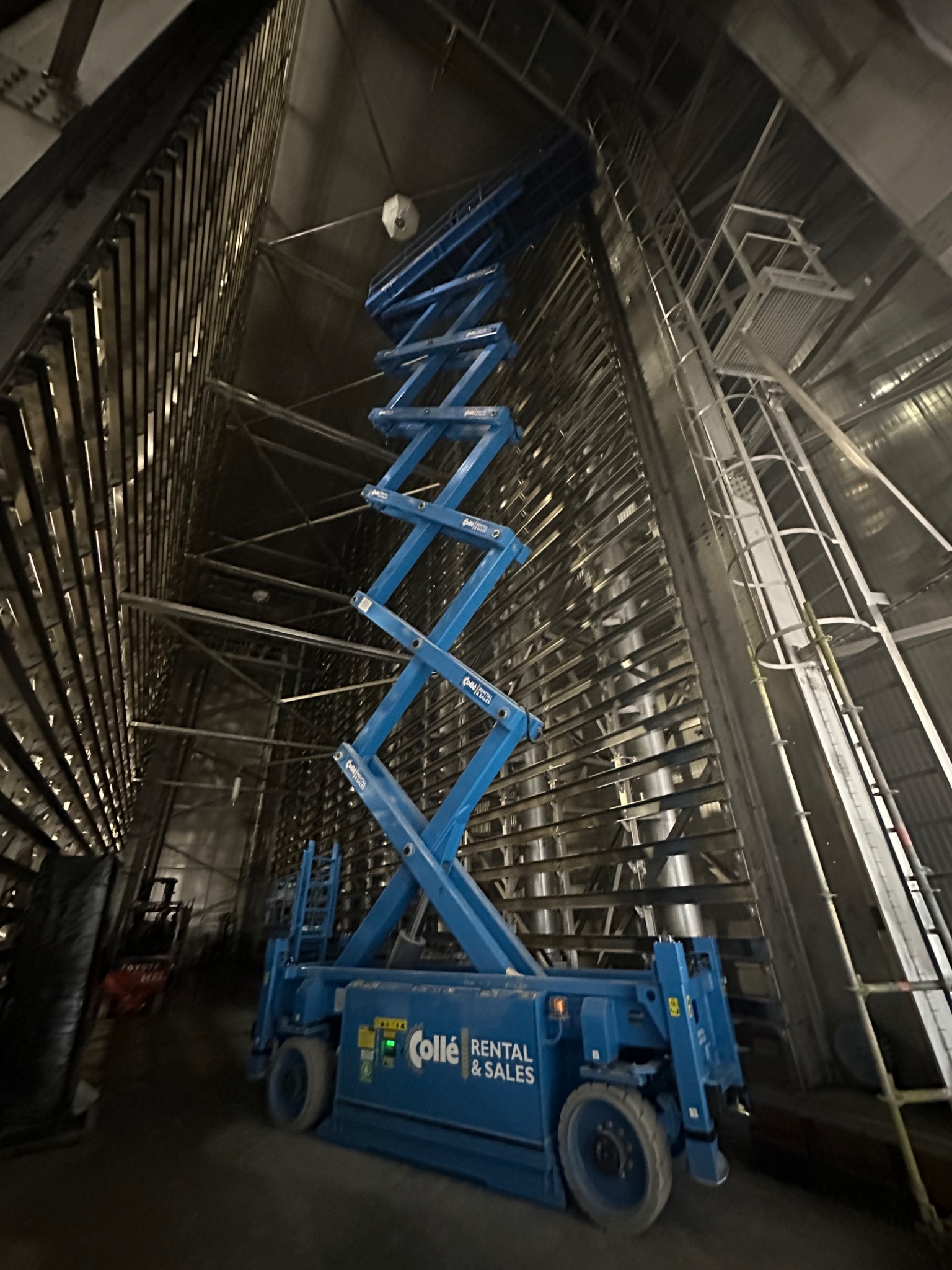 Wnętrze hali przemysłowej z metalową konstrukcją i podnośnikiem nożycowym marki Collé Rental & Sales, widok z dołu ku górze.